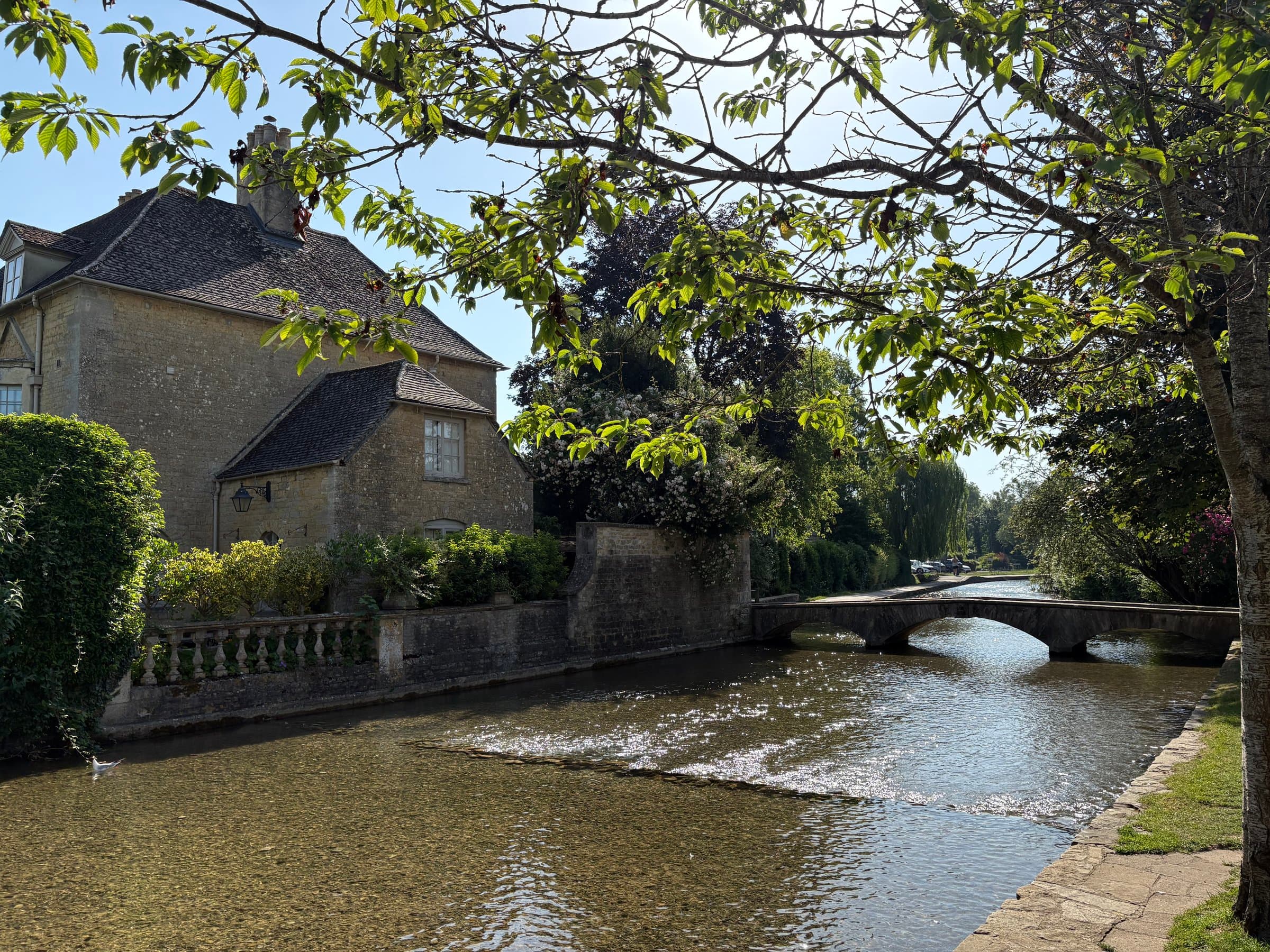 A honey-stone riverside cottage on the Windrush, with one of Bourton's low arched footbridges beyond